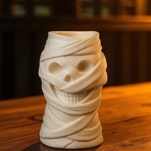 May include: A white ceramic tiki mug shaped like a mummy's head. The mug features a skull face with wrapped bandages, perfect for Halloween or themed parties. The mug is approximately 6 inches tall.