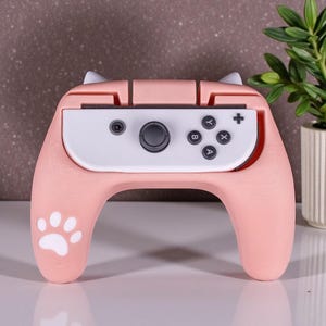 May include: A pink gaming controller with a cat ear design. The controller has a white center section with black buttons and a directional pad. A paw print is on the handle. The controller is designed for a gaming console.