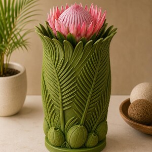 May include: A green vase with a detailed leaf design, holding a pink protea flower. The vase has a textured surface and a wide opening. The flower is a vibrant pink with a spiky center and green leaves.