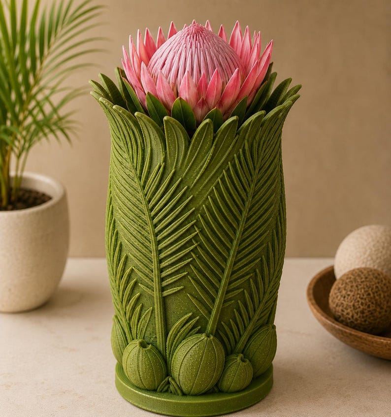May include: A green vase with a detailed leaf design, holding a pink protea flower. The vase has a textured surface and a wide opening. The flower is a vibrant pink with a spiky center and green leaves.