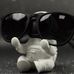 May include: A white astronaut figurine holding a pair of black sunglasses. The sunglasses are resting on the astronaut's head. The figurine is on a dark, speckled surface. The astronaut is in a seated position.