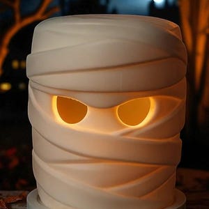 K&ouml;nnte beinhalten: Eine wei&szlig;e, keramikartige Mumien-Halloween-Dekoration mit leuchtend orangefarbenen Augen. Die Mumie ist in strukturierte Bandagen gewickelt und steht auf einem hellblauen Sockel. Der Hintergrund zeigt eine gruselige Nachtszene mit Vollmond.