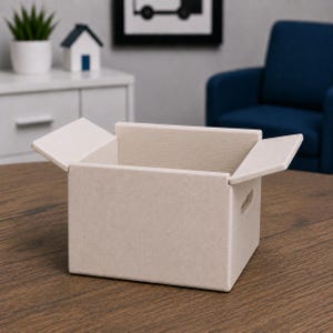 May include: An open, beige cardboard box with two flaps partially open. The box has small cut-out handles on the sides. The box is sitting on a wooden surface. The background includes a white cabinet, a blue armchair, and a framed picture.