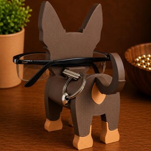 May include: A brown dog-shaped eyeglasses holder with a key ring. The holder has a textured surface and tan accents on the paws. Black eyeglasses rest in the holder. The holder is made of wood and sits on a wooden surface.