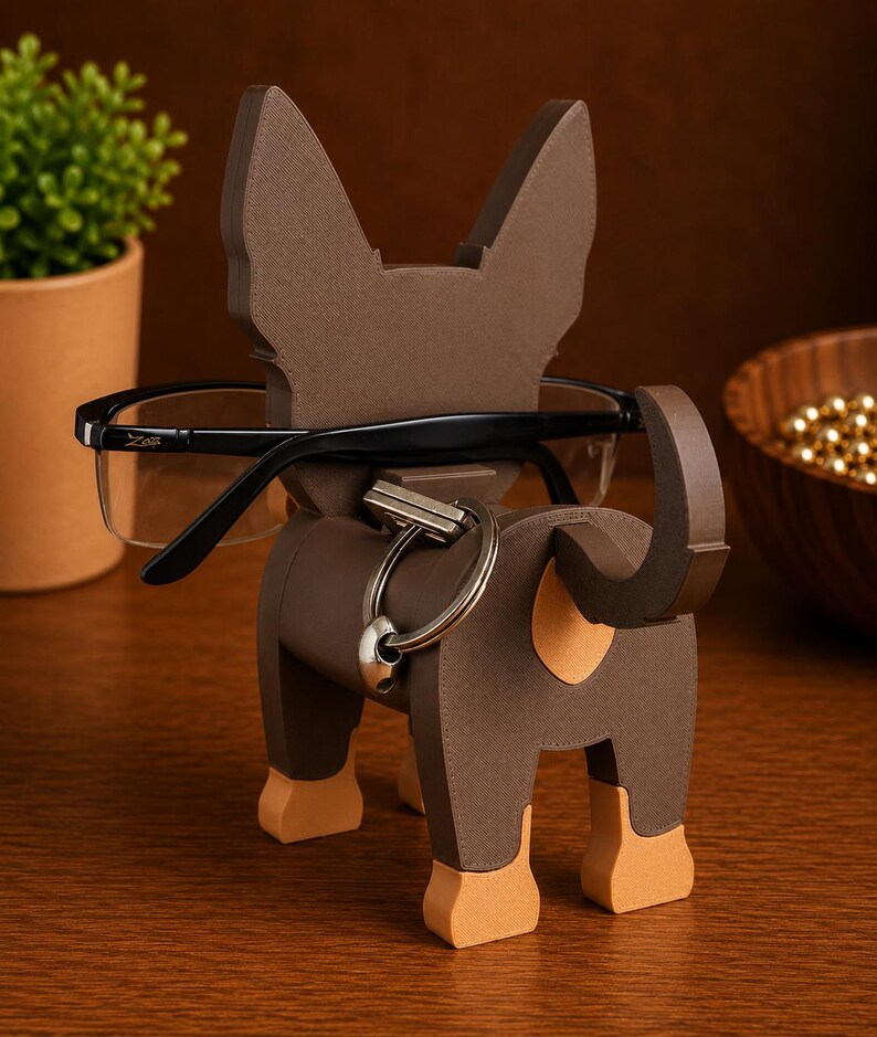 May include: A brown dog-shaped eyeglasses holder with a key ring. The holder has a textured surface and tan accents on the paws. Black eyeglasses rest in the holder. The holder is made of wood and sits on a wooden surface.