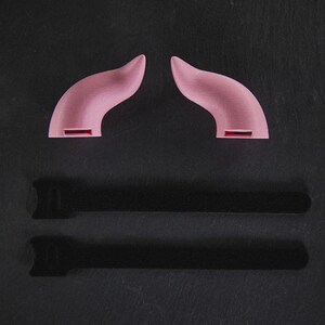May include: Two pink plastic devil horns on a black background. The horns are curved and have a pointed tip. There are two black shadows below the horns.