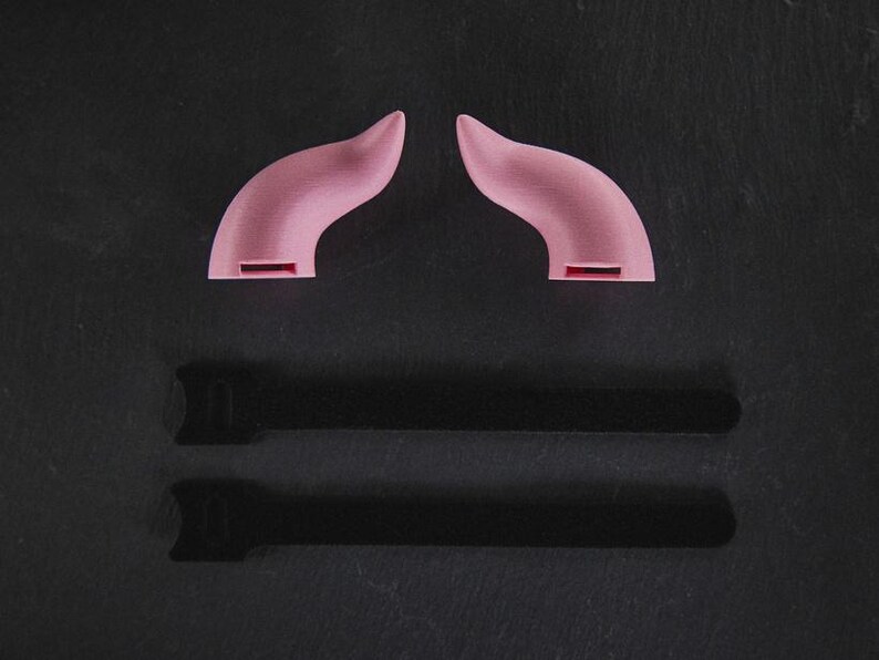 May include: Two pink plastic devil horns on a black background. The horns are curved and have a pointed tip. There are two black shadows below the horns.