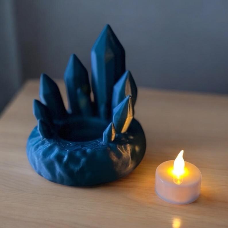 May include: A teal-colored crystal-shaped candle holder with a round base and multiple crystal points. A small, white, battery-operated candle with a flickering yellow light sits next to the holder. The candle holder is on a wooden surface.