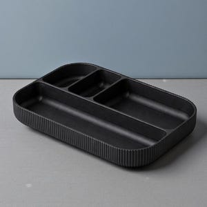 May include: A black rectangular organizer with multiple compartments. The organizer has a ribbed exterior and a smooth interior. The compartments are of varying sizes, designed for organizing small items. The overall design is minimalist and modern.