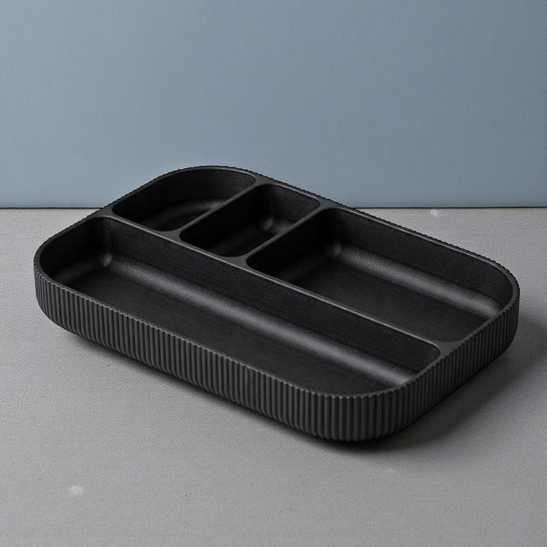 May include: A black rectangular organizer with multiple compartments. The organizer has a ribbed exterior and a smooth interior. The compartments are of varying sizes, designed for organizing small items. The overall design is minimalist and modern.