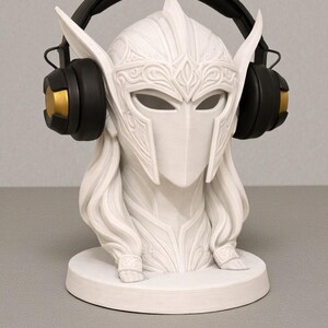 May include: A white bust of a warrior with a helmet and long hair, featuring detailed carvings. Black and gold headphones are placed on the head. The bust sits on a round base, a decorative item.