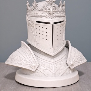 May include: A white decorative bust of a knight's helmet and armor. The helmet features a crown and a visor with small holes. The armor includes shoulder plates and a chest piece, all with detailed relief patterns. The bust is on a round base.