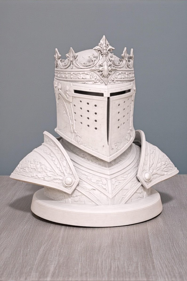 May include: A white decorative bust of a knight's helmet and armor. The helmet features a crown and a visor with small holes. The armor includes shoulder plates and a chest piece, all with detailed relief patterns. The bust is on a round base.