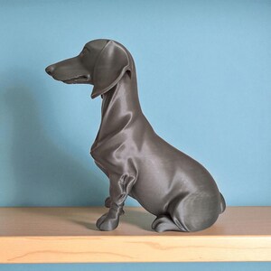 May include: A gray dachshund sculpture sitting on a wooden shelf against a blue wall. The dog is in a seated position, with its head and long snout facing left. The sculpture has a smooth, matte finish.