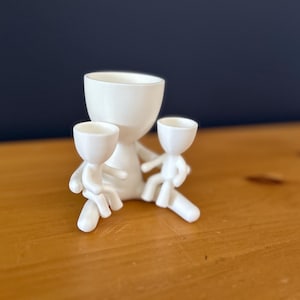May include: A white ceramic planter shaped like a figure, holding two smaller figures. The figures have cup-shaped heads and are sitting. The planter is on a wooden surface with a dark blue background.