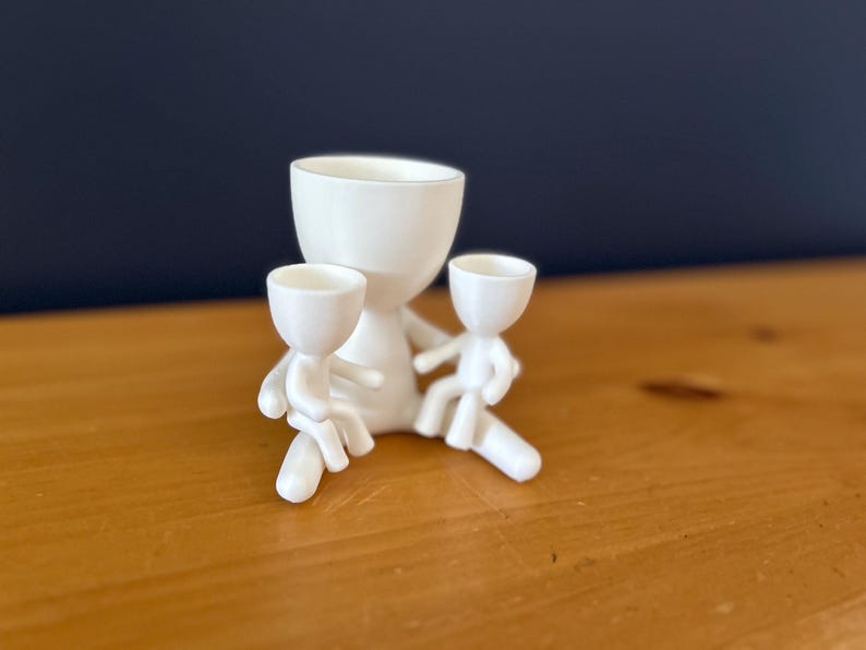 May include: A white ceramic planter shaped like a figure, holding two smaller figures. The figures have cup-shaped heads and are sitting. The planter is on a wooden surface with a dark blue background.