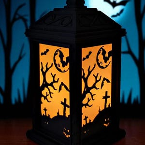 May include: Black Halloween lantern with orange interior light. The lantern features cut-out designs of bats, a full moon, bare trees, crosses, and jack-o'-lantern faces. The background shows a blue sky with a full moon and silhouettes of trees.