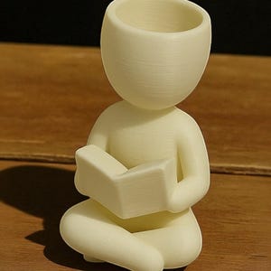 Puede incluir: Figura de color crema de una persona sentada con las piernas cruzadas leyendo un libro. La figura tiene una cabeza redondeada con una parte superior ahuecada, posiblemente para una peque&ntilde;a planta. La figura est&aacute; hecha de un material texturizado.