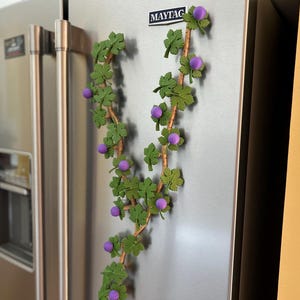 May include: A decorative vine made of green leaves and purple berries is attached to a stainless steel refrigerator. The vine is made of a brown stem with green leaves and purple berries. The refrigerator has a Maytag logo.