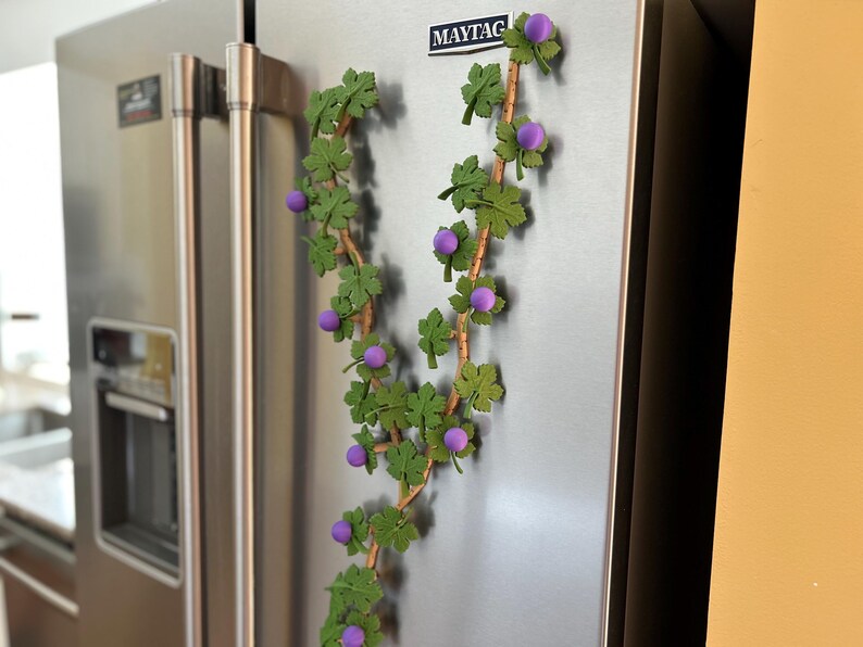 May include: A decorative vine made of green leaves and purple berries is attached to a stainless steel refrigerator. The vine is made of a brown stem with green leaves and purple berries. The refrigerator has a Maytag logo.