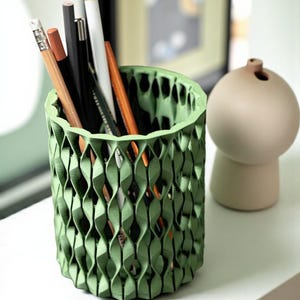 May include: A green, textured pencil holder with a geometric design, filled with pencils and pens of various colors. A small, beige vase is to the right of the holder. The items are on a white surface.