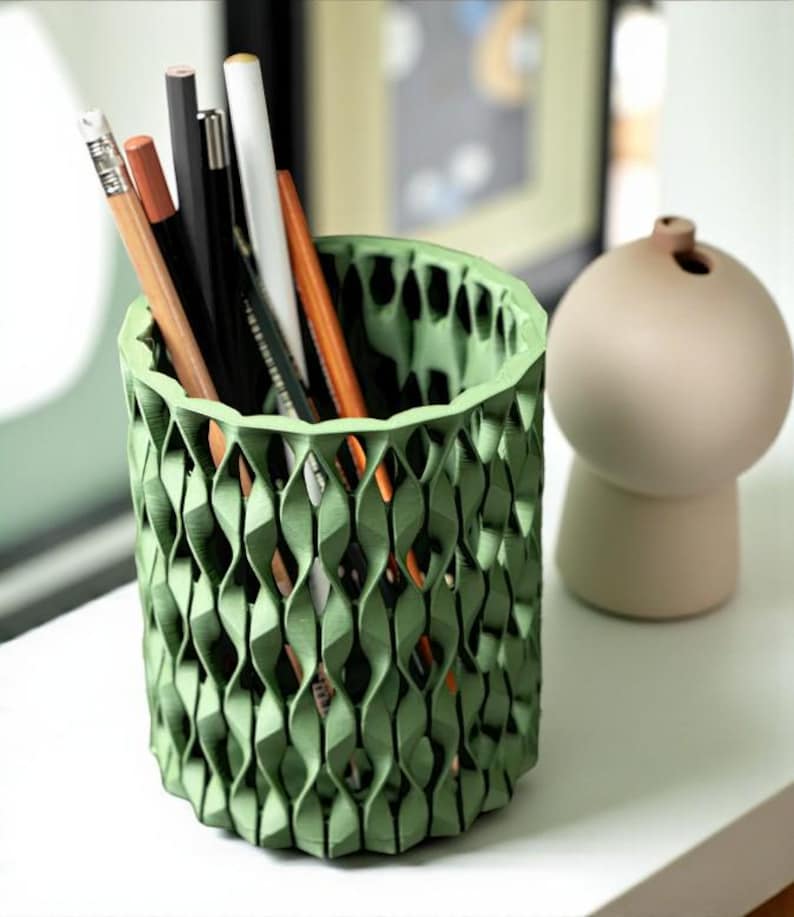 May include: A green, textured pencil holder with a geometric design, filled with pencils and pens of various colors. A small, beige vase is to the right of the holder. The items are on a white surface.