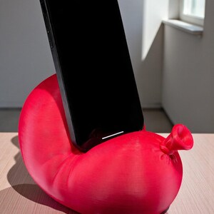 May include: A bright red phone stand shaped like a balloon, with a black smartphone resting in the cradle. The stand has a textured surface and a nozzle detail. The phone is positioned vertically.
