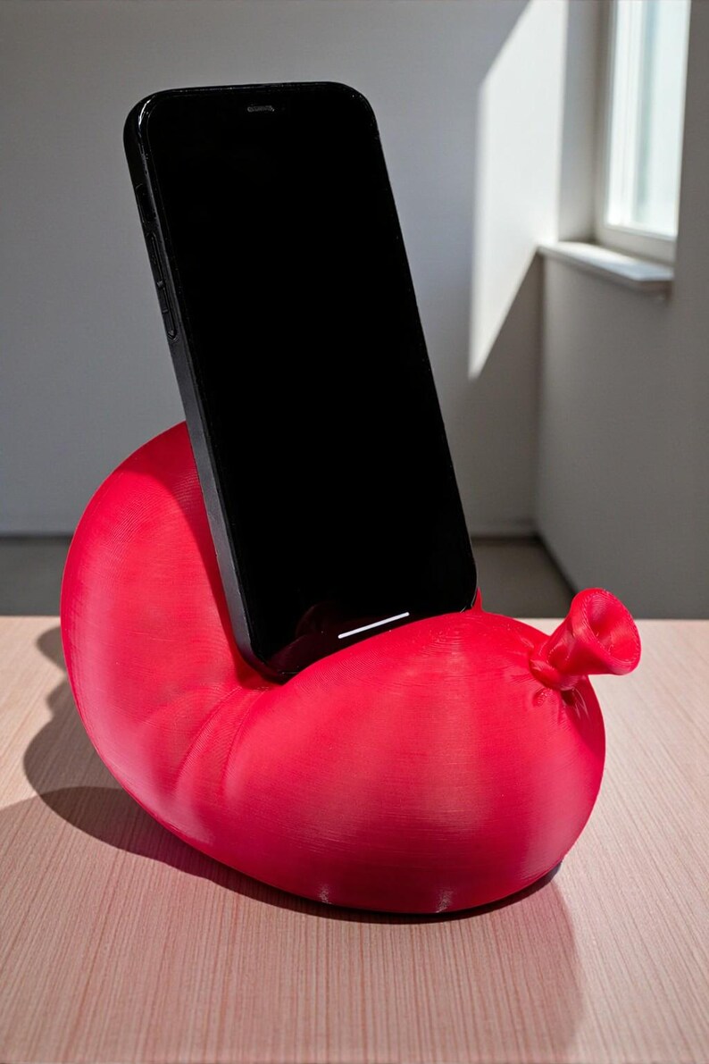 May include: A bright red phone stand shaped like a balloon, with a black smartphone resting in the cradle. The stand has a textured surface and a nozzle detail. The phone is positioned vertically.