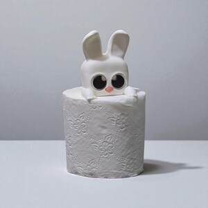 May include: A white bunny figure with large black eyes and a pink nose sits atop a roll of white toilet paper. The toilet paper has a floral embossed pattern. The bunny is a whimsical decorative item.