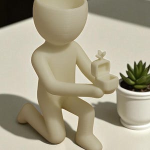 May include: A cream-colored figurine of a person kneeling, holding an open ring box with a butterfly on top. The figurine has a large, open head, suitable for a small plant. A small potted succulent is to the right.