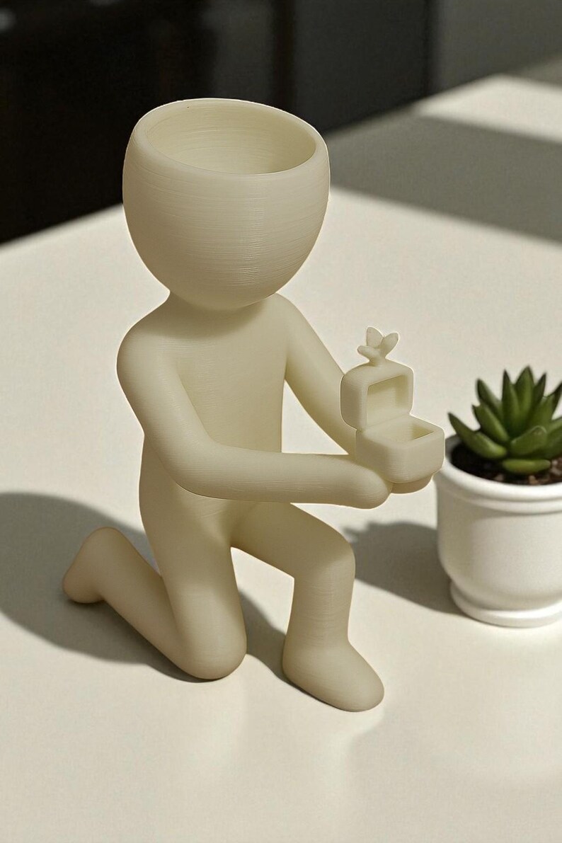May include: A cream-colored figurine of a person kneeling, holding an open ring box with a butterfly on top. The figurine has a large, open head, suitable for a small plant. A small potted succulent is to the right.