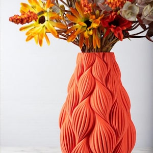 May include: An orange vase with a textured, leaf-like pattern holds a bouquet of colorful flowers. The flowers include yellow, orange, and red blooms, with green and brown foliage. The vase is a vibrant accent piece.