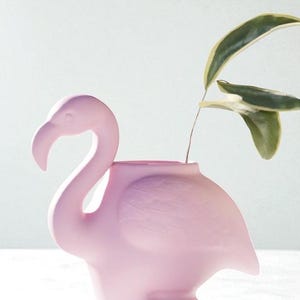 Puede incluir: Un jarrón rosa claro con forma de flamenco, con un cuello largo y curvo y un pico. El jarrón contiene un tallo de planta verde y amarilla. El flamenco tiene un acabado liso y mate y es un elemento decorativo.