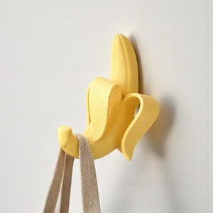 May include: A yellow banana-shaped wall hook with a beige tote bag hanging from it. The hook is mounted on a white wall. The banana peel is partially open, revealing the hook.