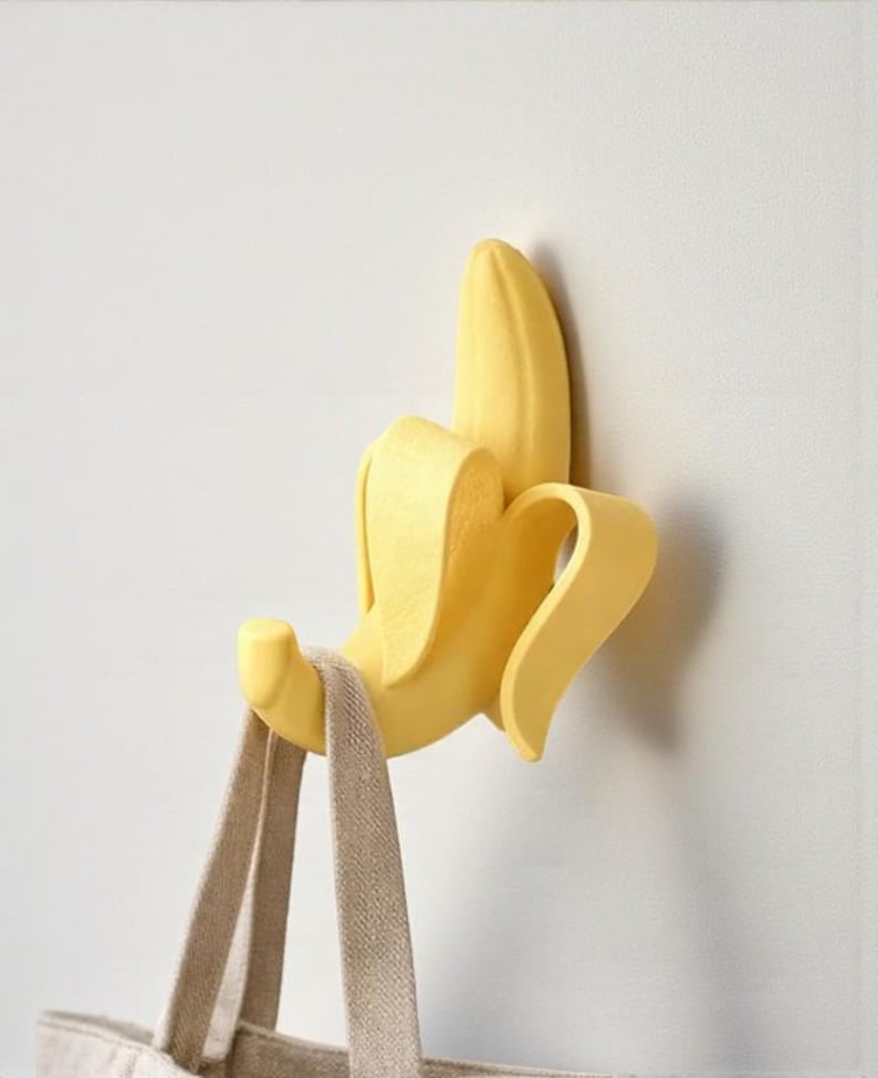 May include: A yellow banana-shaped wall hook with a beige tote bag hanging from it. The hook is mounted on a white wall. The banana peel is partially open, revealing the hook.