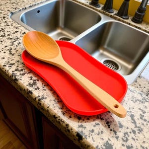 Puede incluir: Una cuchara de madera descansa sobre un reposacucharas de silicona rojo brillante. La cuchara es de color marr&oacute;n claro con un cuenco redondeado y un mango largo. El reposacucharas es rectangular con bordes curvos. El fregadero de la cocina est&aacute; al fondo.