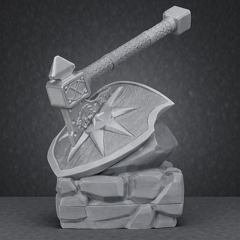 May include: A gray, 3D-printed fantasy axe and shield set. The axe features a textured handle and a hammerhead design. The shield has a starburst pattern and is resting on a stone base, suggesting a miniature display piece.