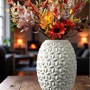 May include: A white, textured vase filled with colorful faux flowers. The vase has a geometric pattern and sits on a wooden table. The flowers include red, orange, and yellow blooms, with various green and brown stems.