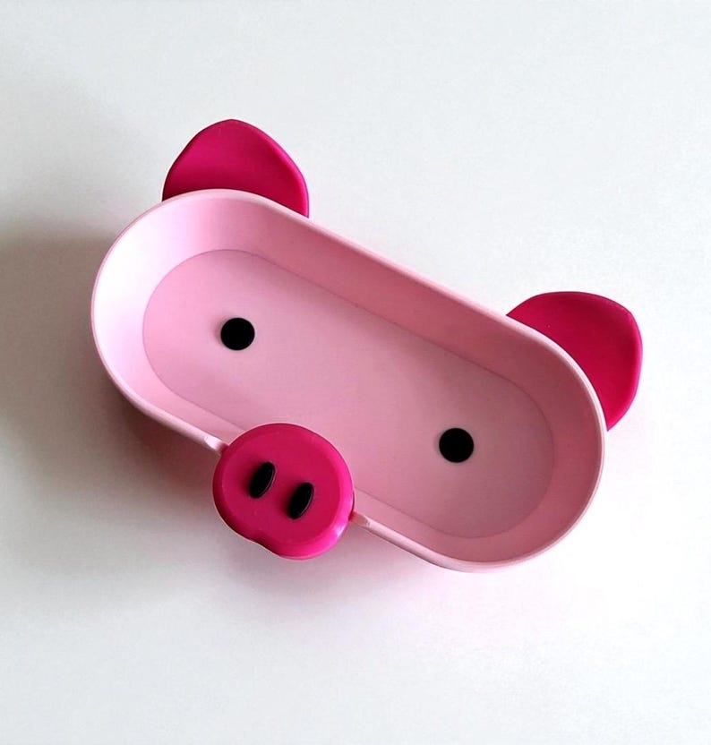 Op de afbeelding: Een roze, ovaalvormige schaal met een varkensontwerp. De schaal heeft twee zwarte stippen voor ogen, een roze snuit met twee zwarte neusgaten en twee roze oren. Het is gemaakt van glad materiaal op een witte achtergrond.