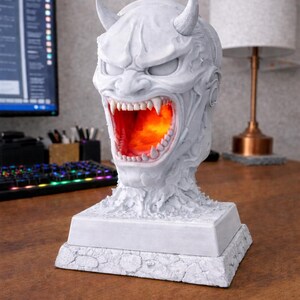 May include: A white, detailed sculpture of a demonic face with horns and an open mouth revealing an orange glow. The sculpture sits on a square base. The piece is a decorative lamp.