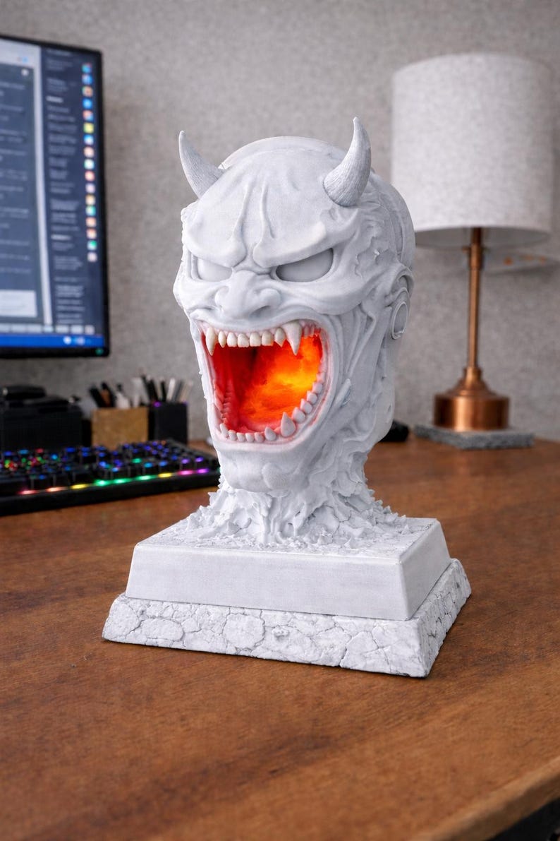 May include: A white, detailed sculpture of a demonic face with horns and an open mouth revealing an orange glow. The sculpture sits on a square base. The piece is a decorative lamp.