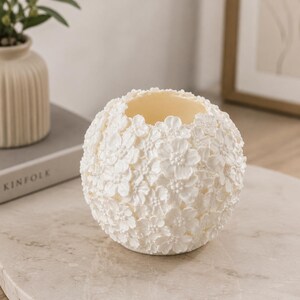 May include: A white, spherical candle holder covered in small, three-dimensional flower blossoms. The interior is a light yellow color. The candle holder is sitting on a marble-like surface.