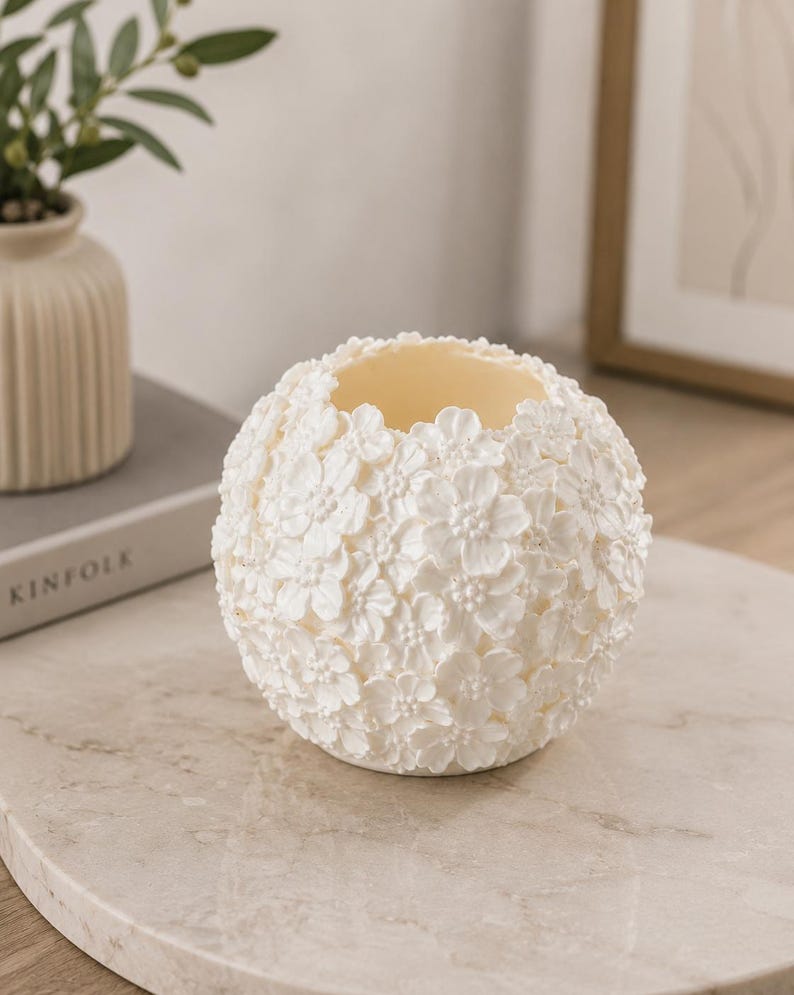 May include: A white, spherical candle holder covered in small, three-dimensional flower blossoms. The interior is a light yellow color. The candle holder is sitting on a marble-like surface.