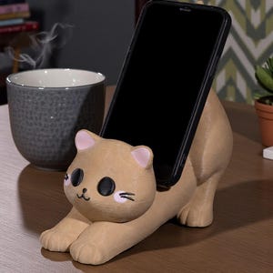 May include: A tan cat-shaped phone holder with a black phone in place. The holder has pink accents on the ears and cheeks, with black eyes and whiskers. The phone holder is on a wooden surface.