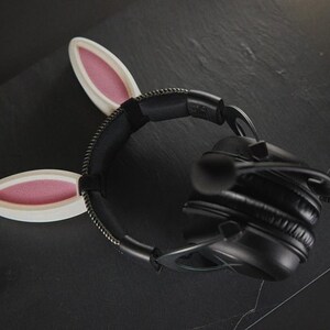 May include: Black headphones with a white headband featuring two pink bunny ears. The headband is made of a soft, flexible material.