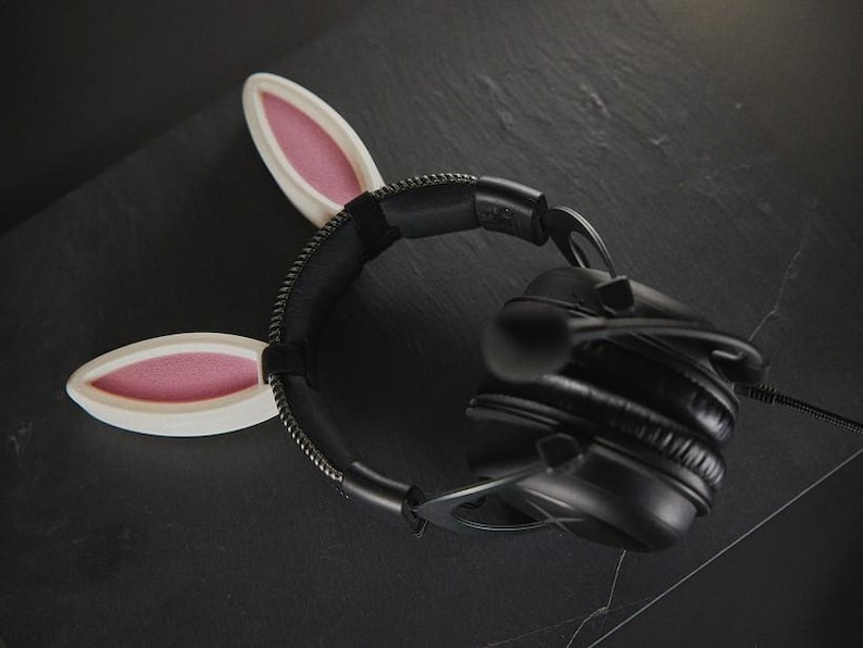 May include: Black headphones with a white headband featuring two pink bunny ears. The headband is made of a soft, flexible material.