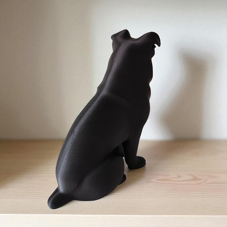 May include: A black dog figurine, viewed from the rear, sitting on a light-colored wooden shelf. The dog's form is smooth and solid, with a slight texture visible. The figurine is a decorative item.