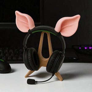 以下が含まれることがあります： 黒いゲーミングヘッドホンとマイク。木製のスタンドに置かれています。ヘッドホンにはピンクの豚の耳が飾られています。スタンドは白い表面にあり、背景には黒いコンピューターマウスが見えます。