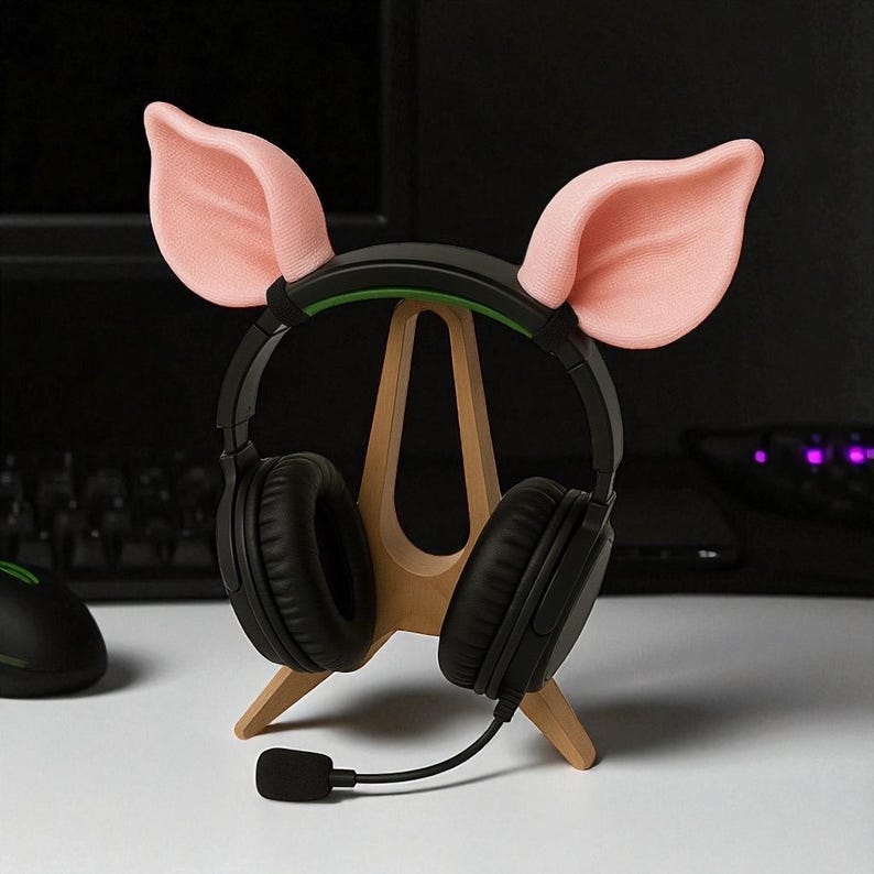 以下が含まれることがあります： 黒いゲーミングヘッドホンとマイク。木製のスタンドに置かれています。ヘッドホンにはピンクの豚の耳が飾られています。スタンドは白い表面にあり、背景には黒いコンピューターマウスが見えます。