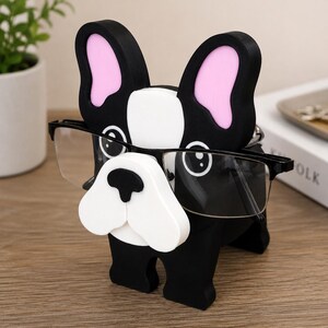 May include: A black and white French Bulldog-shaped eyeglasses holder with pink inner ears. The holder is designed to hold a pair of eyeglasses. The glasses are black-framed and sit in the dog's face.